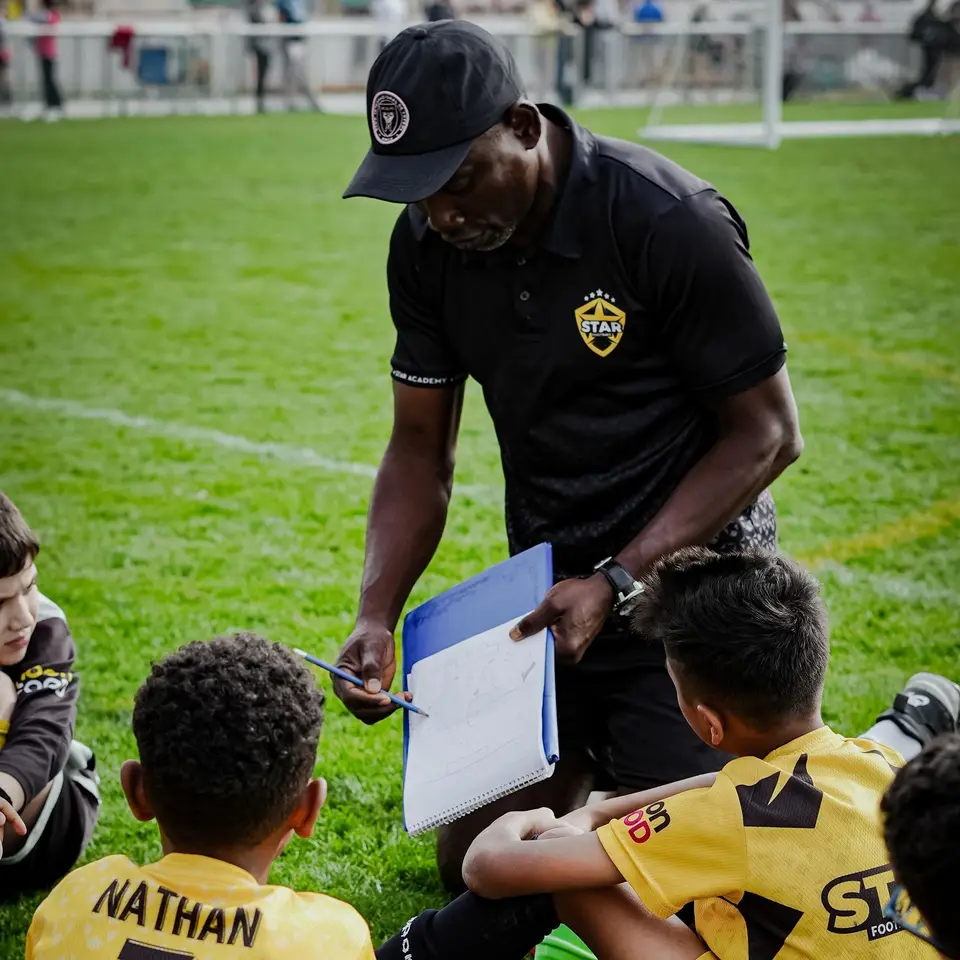 A coach with a clipboard crouched among young players on a pitch