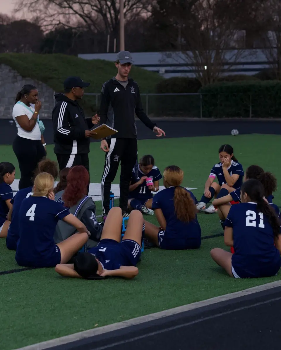 A girls’ team sitting on an artificial pitch while their coaches talk